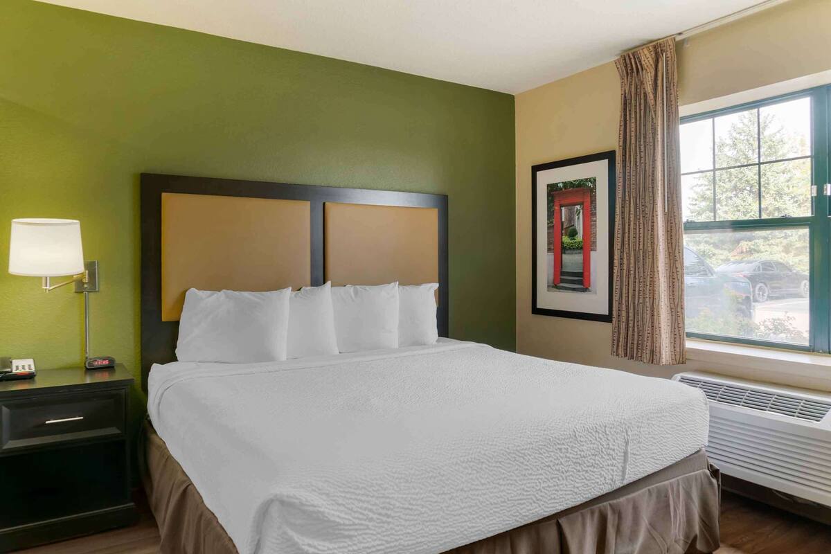 A hotel room with a large bed, a nightstand with a lamp, a framed picture on the wall, and a window with curtains.