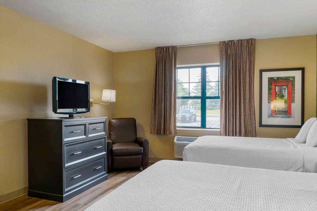 A hotel room with two beds, a TV on a dresser, a chair, and a window with curtains.