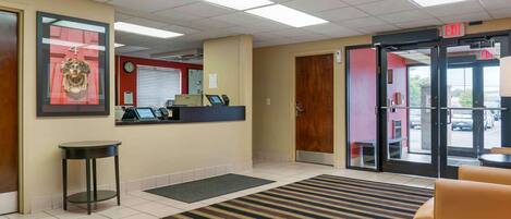 A hotel reception area with a reception desk, a clock, a door, and a waiting area with a striped rug.