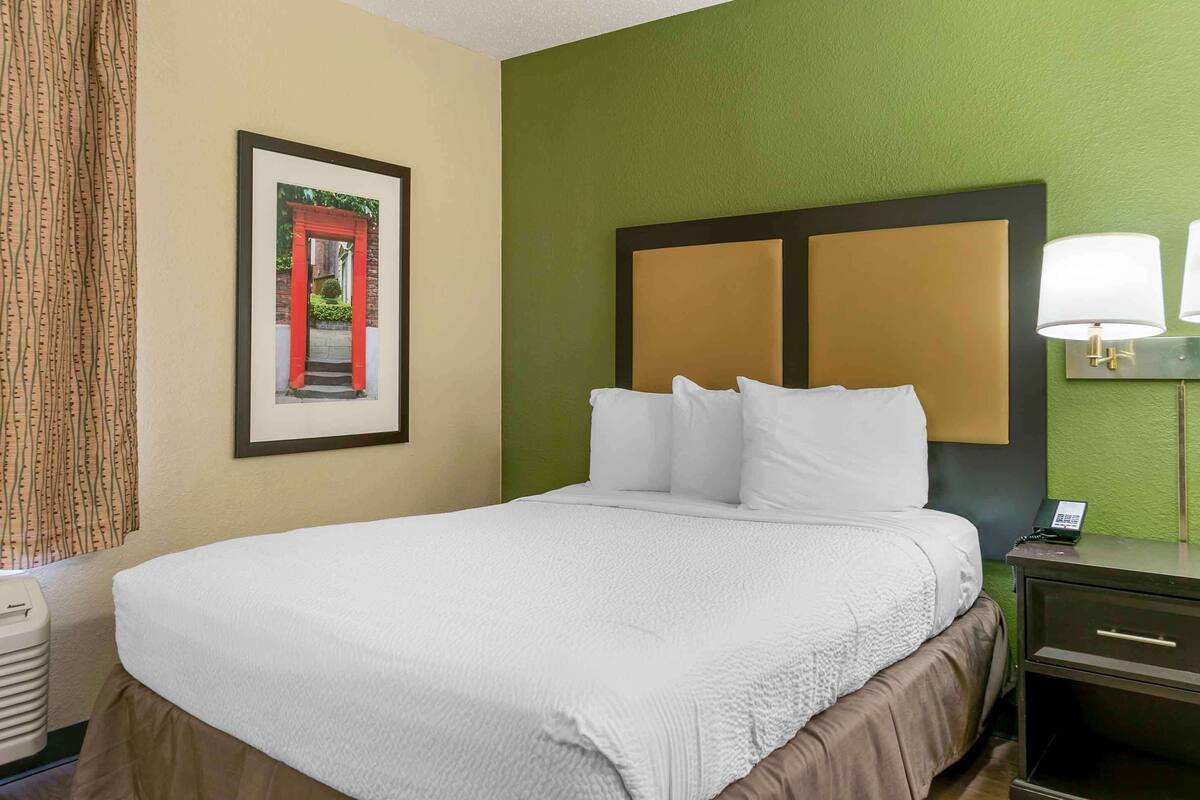 A neatly made bed with white linens, a framed picture of a red gate on the wall, and a bedside lamp.