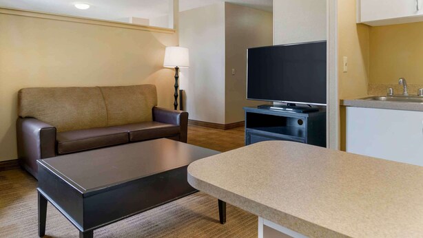 A compact living area with a sofa, a coffee table, a flat-screen TV, a lamp, and a kitchenette with a sink. - Extended Stay America Suites Detroit Madison Heights (Madison Heights)