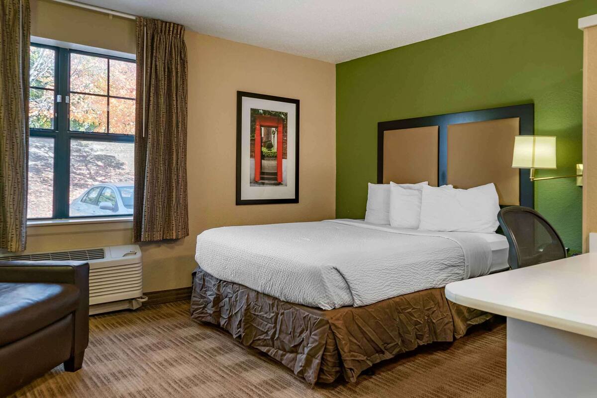 Ein Hotelzimmer mit einem großen Bett, einem Schreibtisch, einem Sessel, einem gerahmten Bild an der Wand und einem Fenster mit Vorhängen.