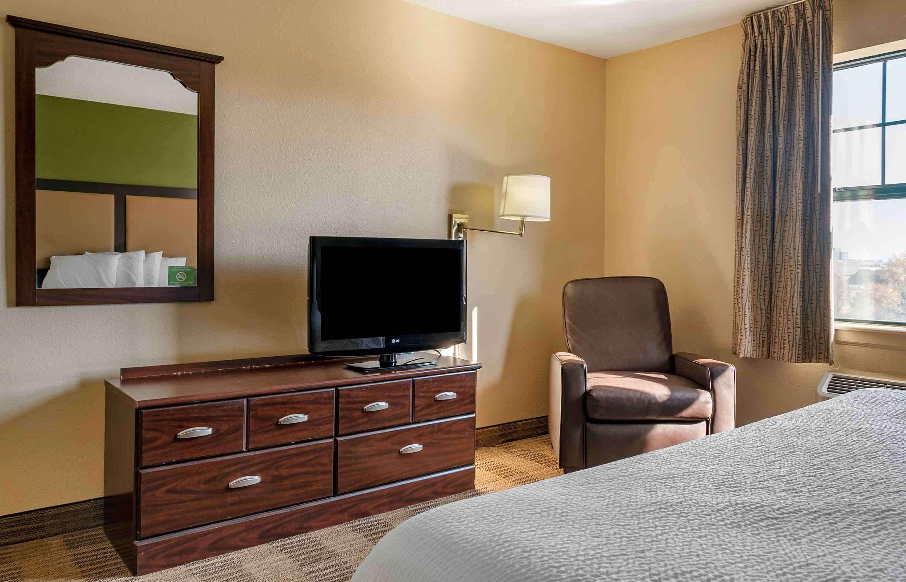 Habitación de hotel con una cama, un televisor sobre un aparador de madera, una silla y una ventana con cortinas.
