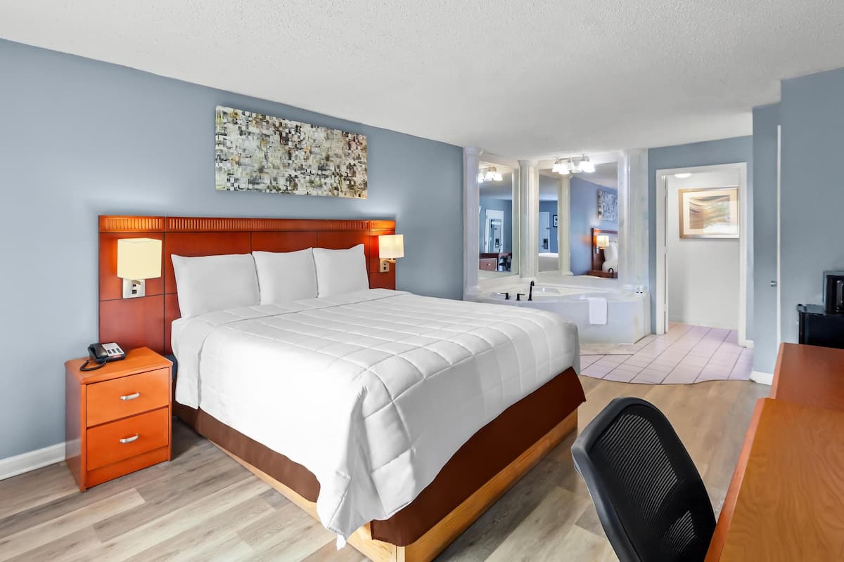 Habitación de hotel con cama, mesita de noche, escritorio, silla y baño con bañera y espejo.