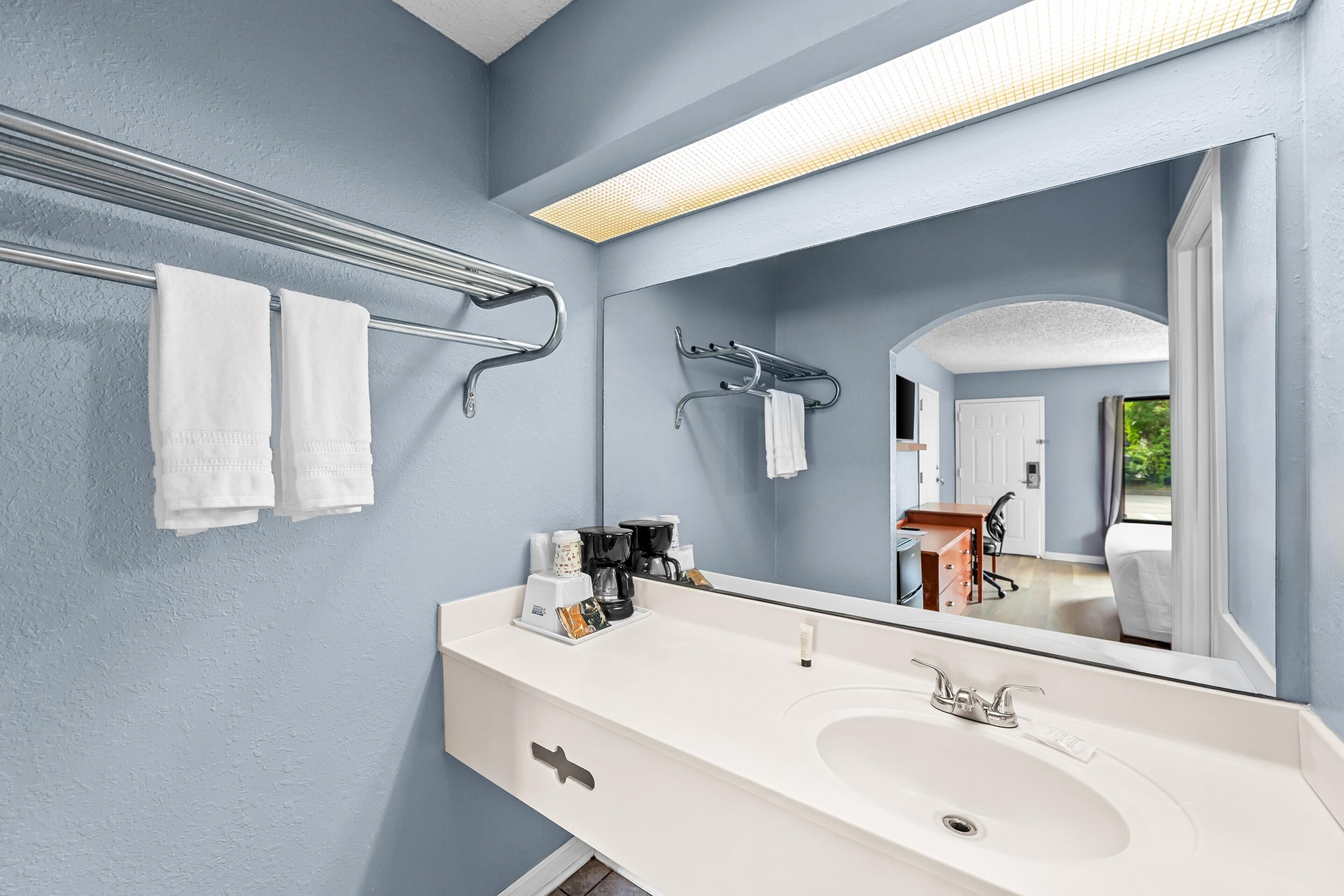 A modern bathroom with a large mirror, white sink, towel rack, and coffee maker.