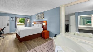 A bedroom with a bed, bedside table, lamp, and a large mirror reflecting a bathroom with a bathtub. - Americas Best Value Inn Brownsville West Tennessee (Brownsville)