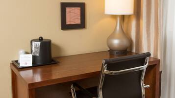 A hotel desk with a black office chair, a lamp, a coffee maker, and two framed pictures on the wall.