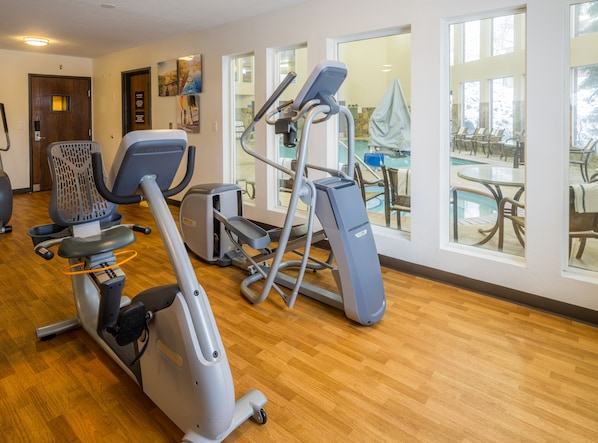 Un gimnasio con máquinas de cardio, vista a la piscina y suelo de madera.