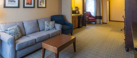 A hotel room with a grey sofa, a wooden coffee table, a blue armchair, a kitchenette, a red chair, and a door leading to another room.