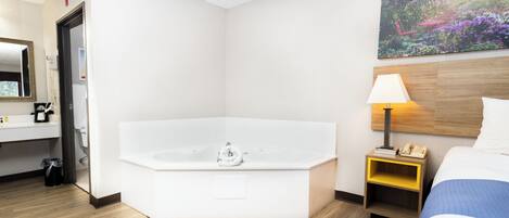 Habitación de hotel con una bañera grande, una mesita de noche con lámpara, una cama, un cuadro en la pared y un baño con lavabo y espejo.