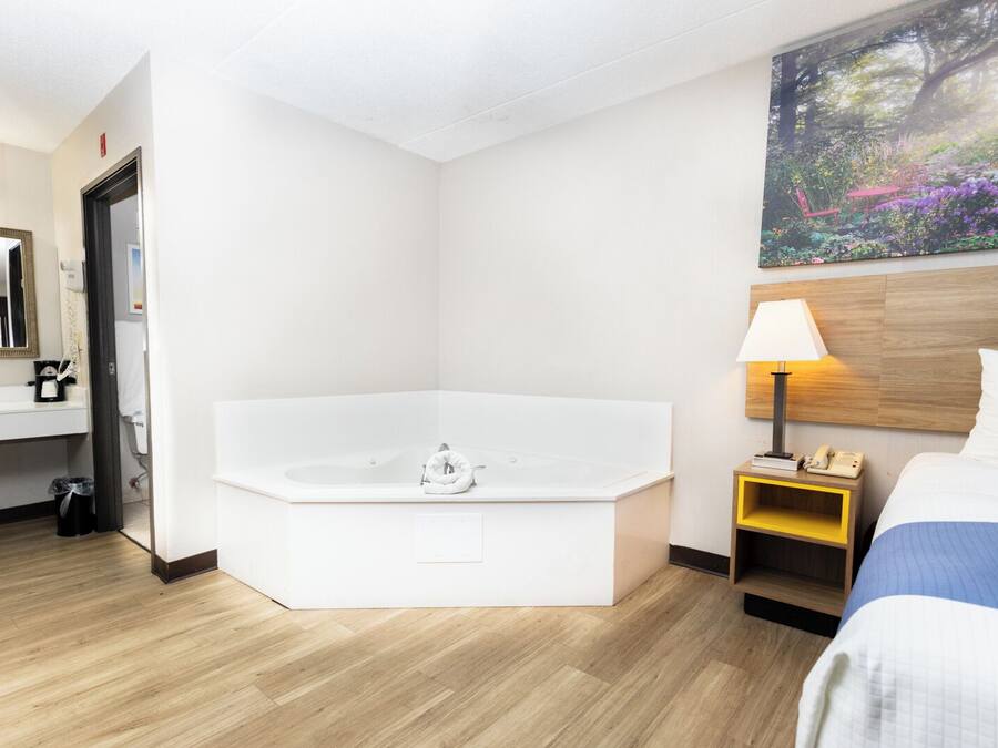 Habitación de hotel con una bañera grande, una mesita de noche con lámpara, una cama, un cuadro en la pared y un baño con lavabo y espejo.