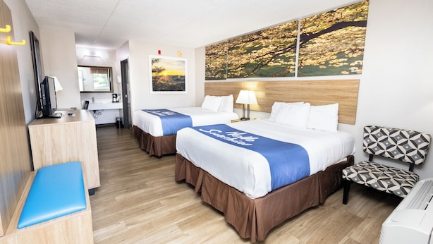 Quarto de hotel com duas camas, uma escrivaninha com computador, uma cadeira e uma janela grande com vista para árvores.