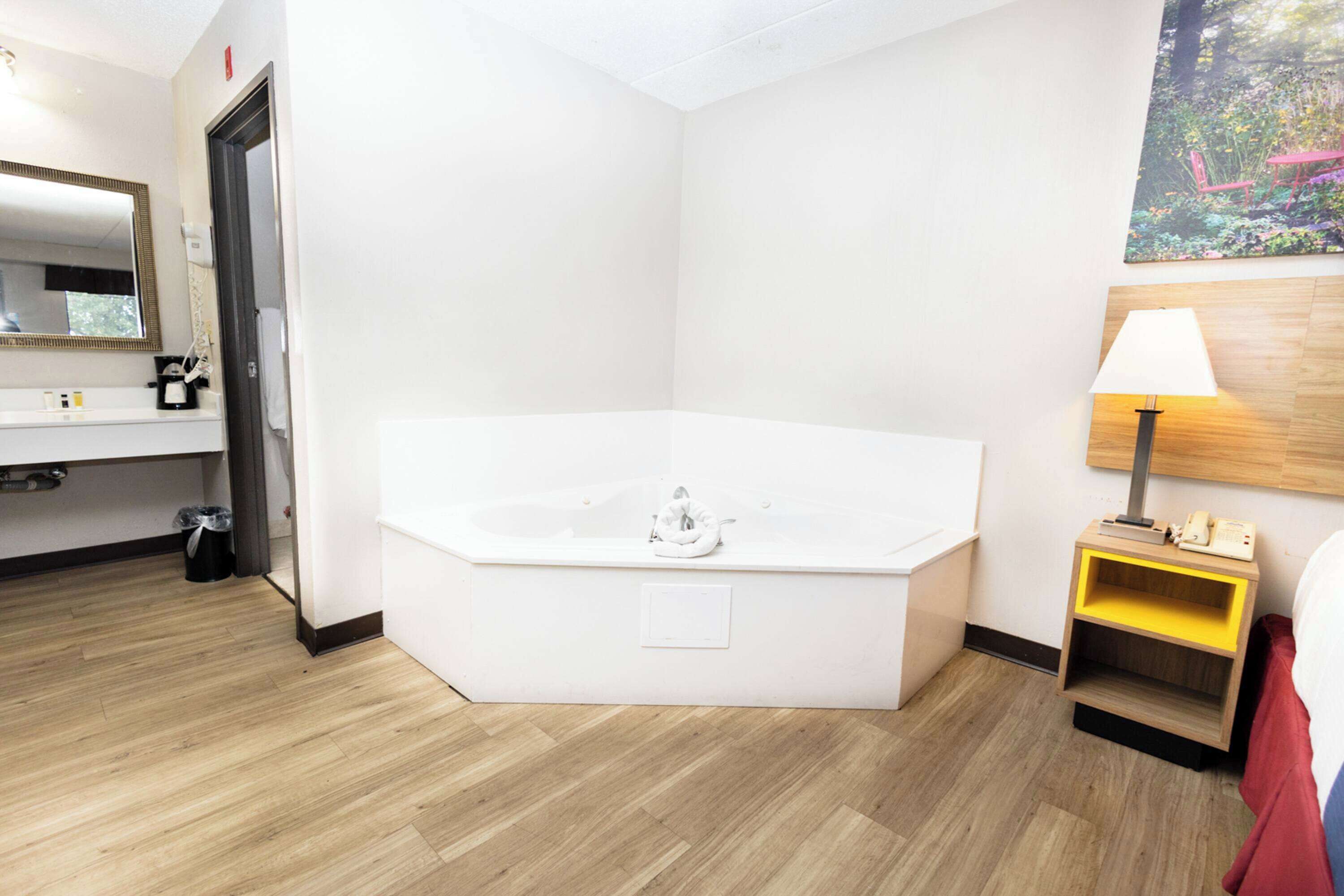 A modern hotel room with a large bathtub, a wooden floor, a bedside table with a lamp, and a painting on the wall.