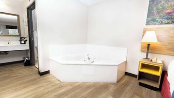 A modern hotel room with a large bathtub, a wooden floor, a bedside table with a lamp, and a painting on the wall.