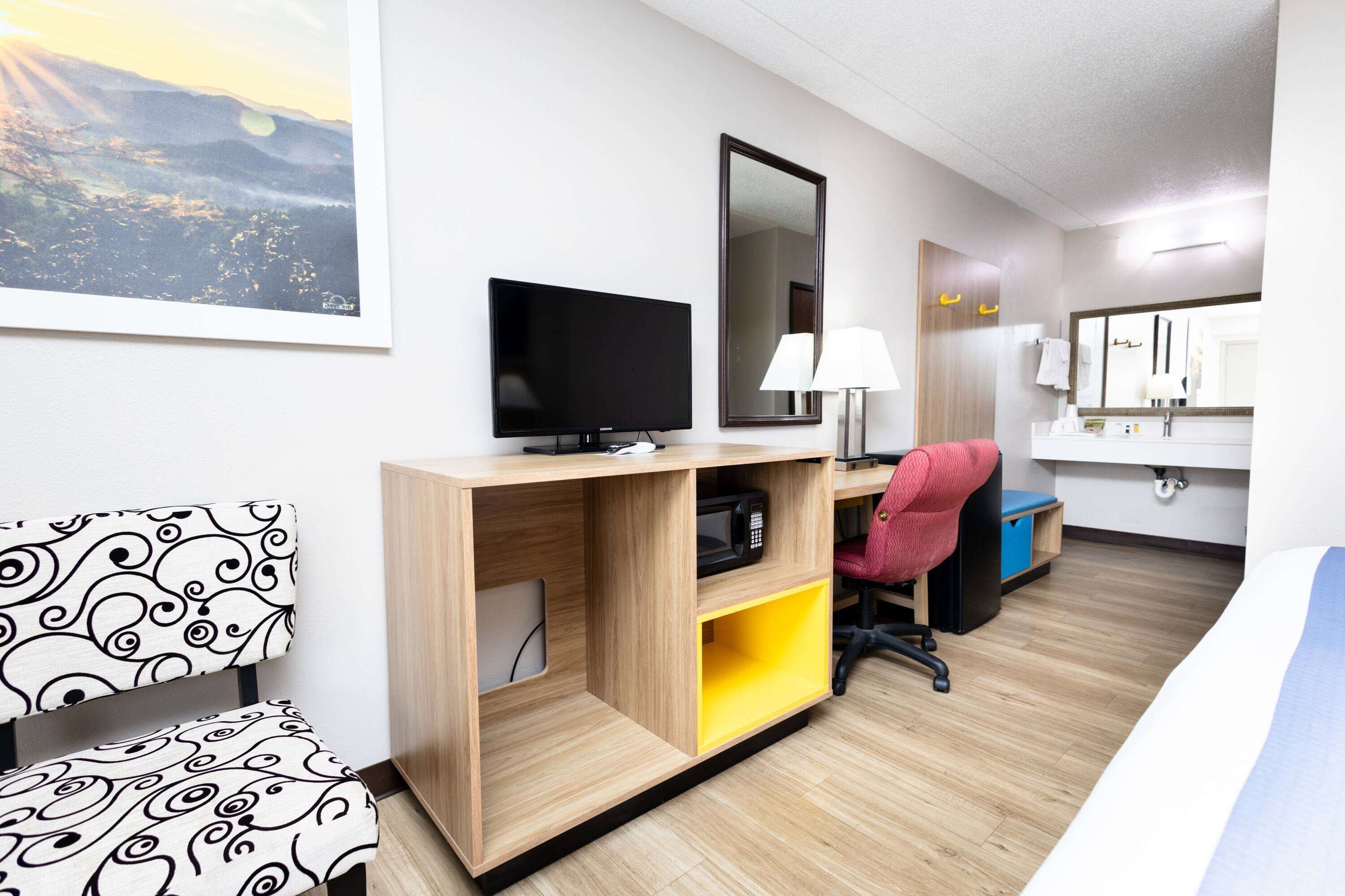 Ein Hotelzimmer mit Bett, Schreibtisch mit Computer, Stuhl, Fernseher, Spiegel und einem Badezimmer mit Waschbecken und Spiegel.