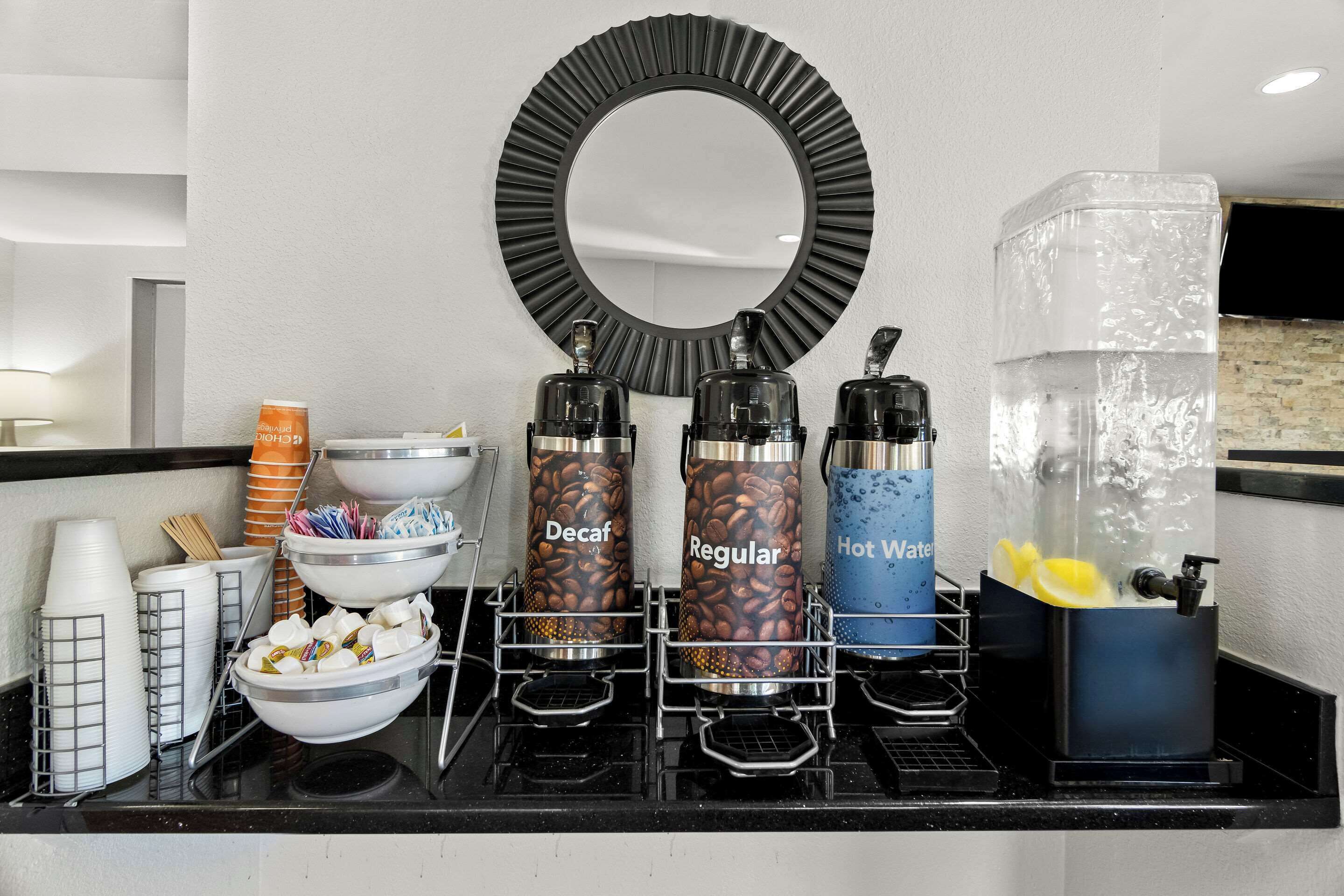 A coffee station with decaf, regular, and hot water dispensers, a large mirror, and various coffee cups and mugs.
