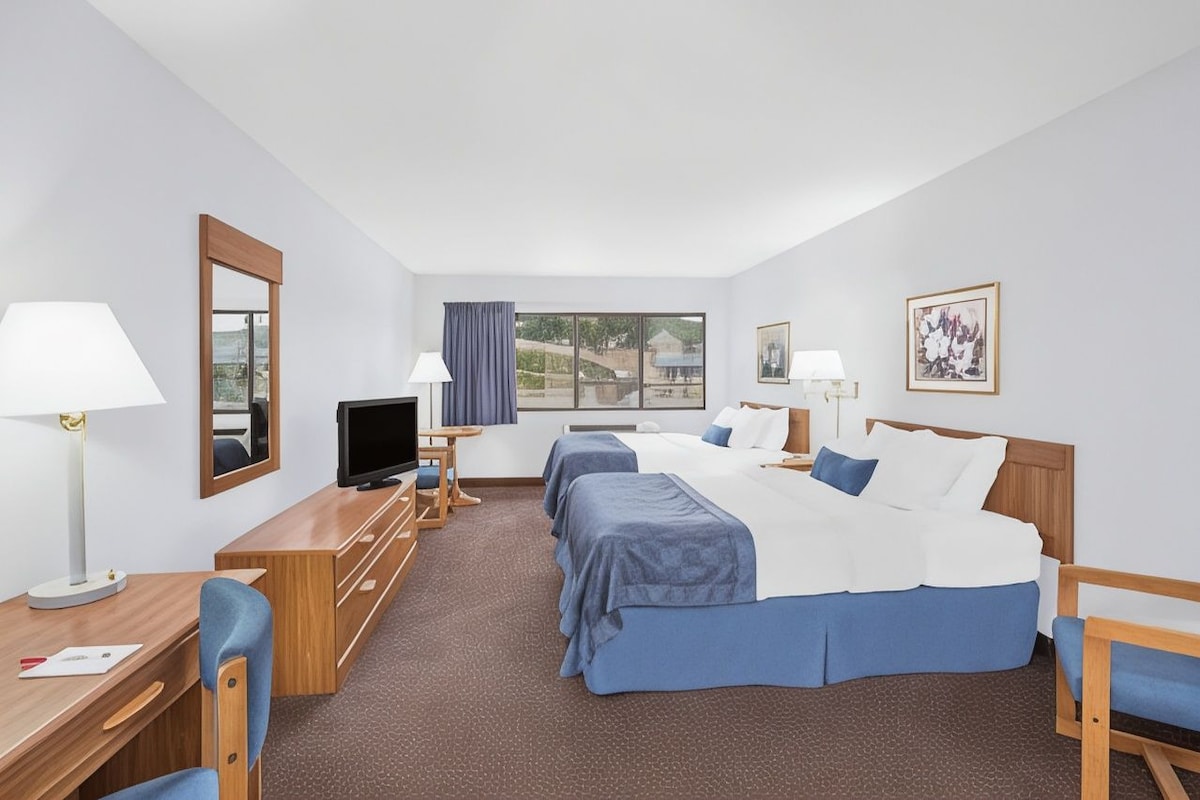 A hotel room with two beds, a desk with a chair, a television, and a window with a view of a bridge.
