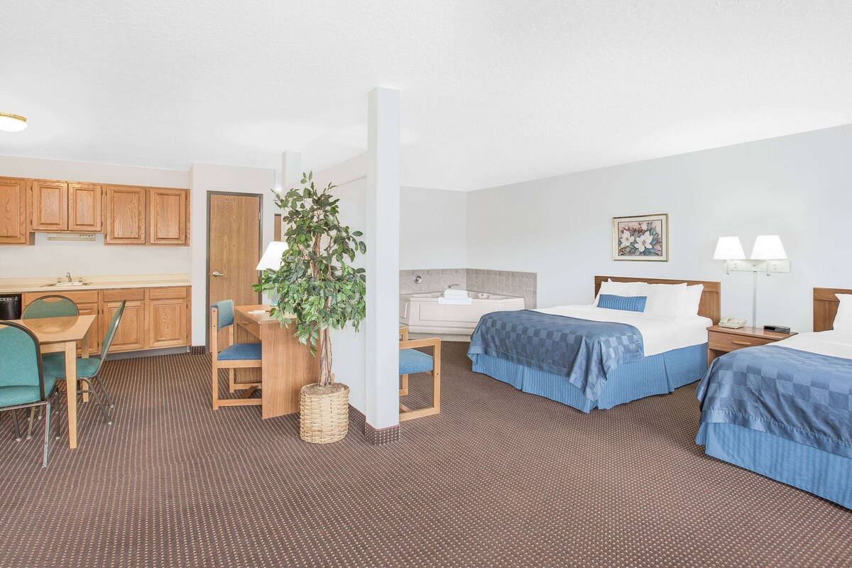 A hotel room with two beds, a desk, a chair, a small table, a plant, a bathroom, and a kitchenette.