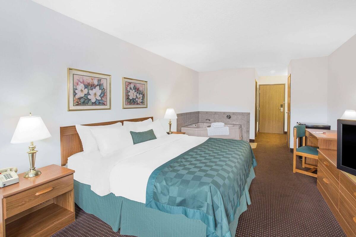 A hotel room with a bed, a desk with a computer, a desk lamp, a telephone, a television, and a bathroom with a bathtub.