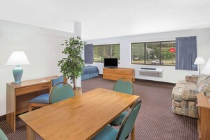 Habitación de hotel con mesa de comedor de madera, sillas azules, un sofá, un televisor y un ventanal con vistas al exterior.