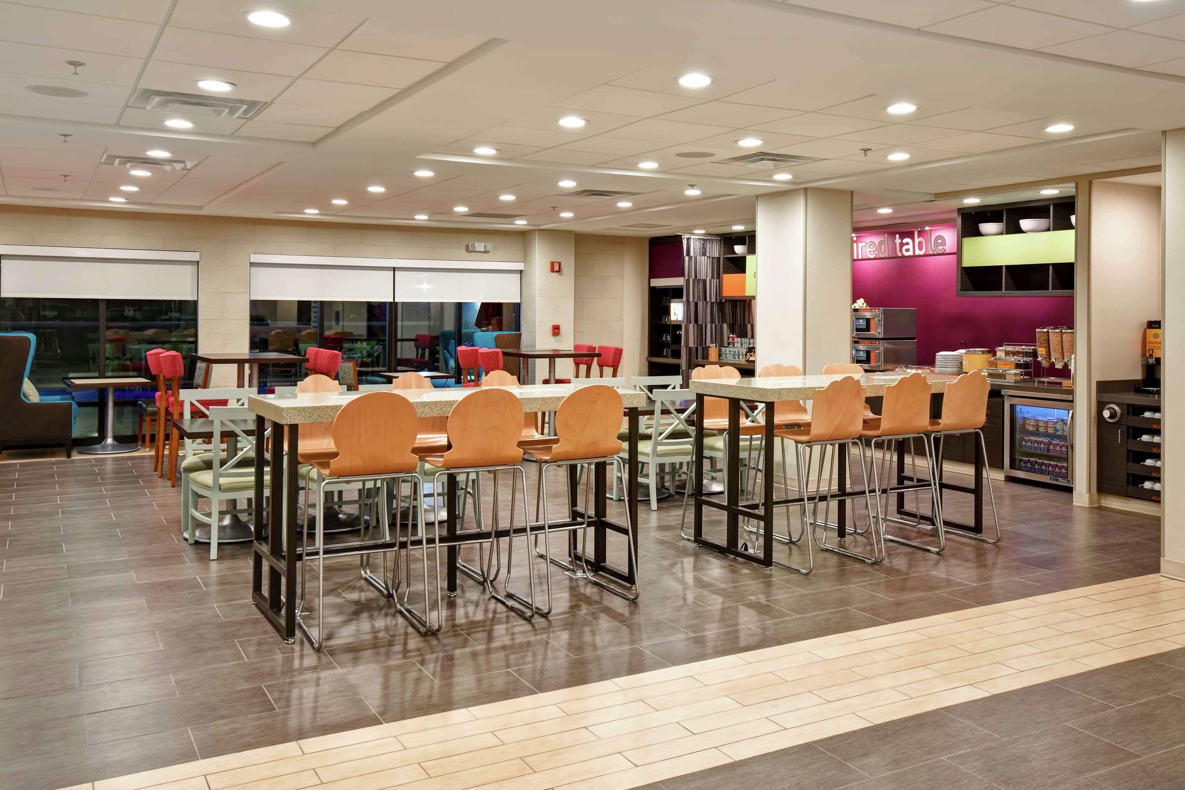 A modern cafeteria with multiple tables and chairs, a vending machine, and a counter with a purple sign.