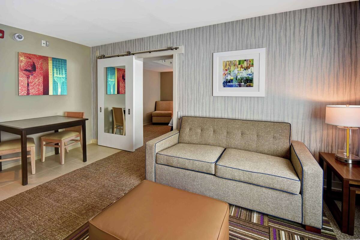 A hotel room with a sofa, a small table, a chair, a lamp, and two framed artworks on the wall.