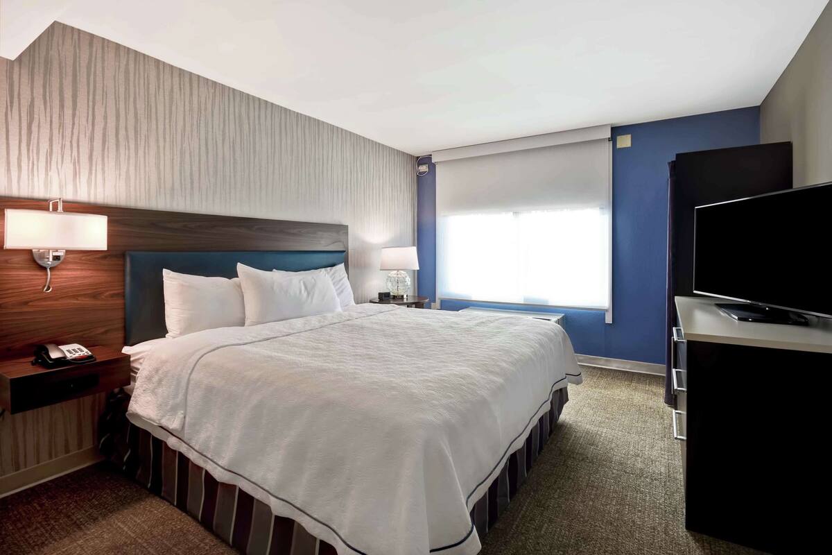A hotel room with a large bed, a desk with a flat-screen TV, a telephone, and a lamp.