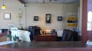 A reception desk with a computer, printer, and various office supplies. There are framed pictures on the wall, a plant, and a calendar.