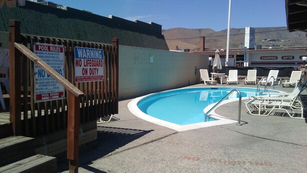 A swimming pool with a wooden fence and multiple signs, including 'NO LIFEGUARD ON DUTY' and 'WARNING NO LIFEGUARD ON DUTY'.