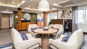 A modern hotel lobby with a round wooden table surrounded by four beige armchairs, a reception desk, and a large abstract painting on the wall. - Fairfield Inn by Marriott St. George (St. George)