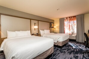 A hotel room with two beds, a large window with curtains, a desk with a lamp, and a chair.
