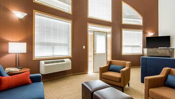 A hotel room with a large window, a flat-screen TV, and a seating area with sofas and ottomans.