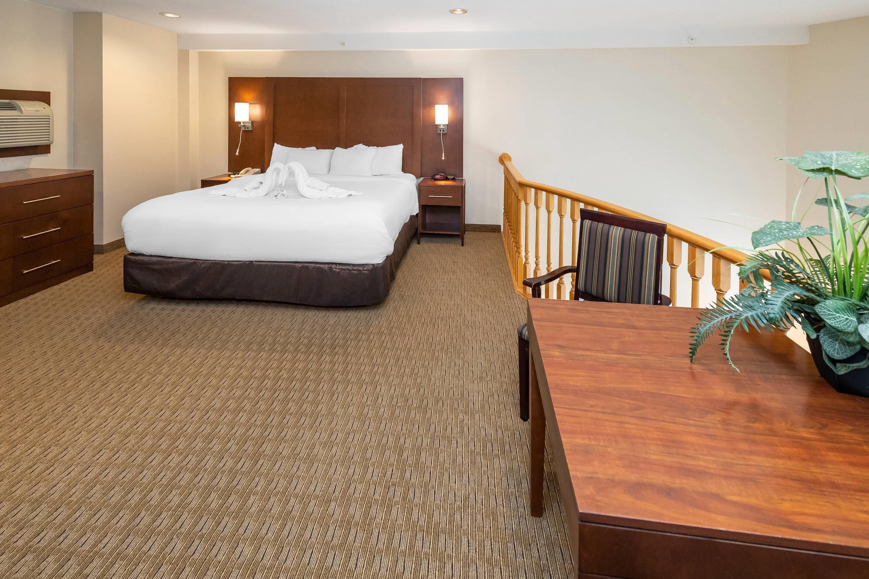 A hotel room with a large bed, a wooden desk, a nightstand, a lamp, and a staircase.