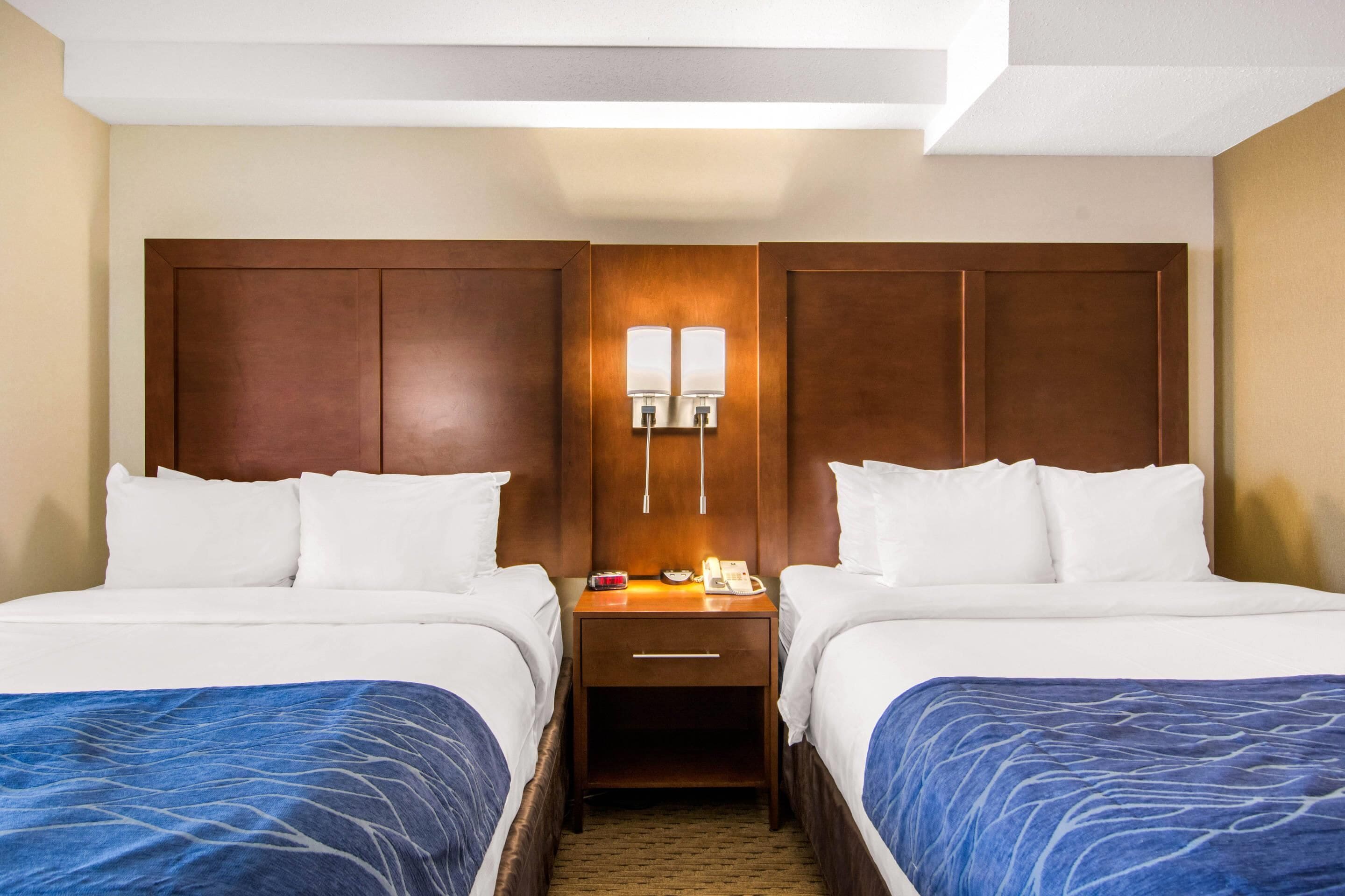 A hotel room with two beds, a nightstand with a phone and a lamp, and a wooden headboard.