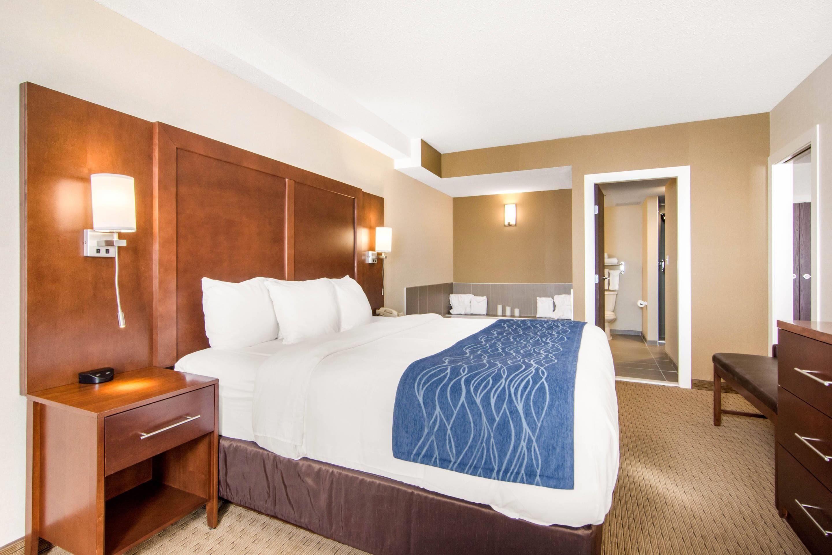 Una habitación de hotel con una cama grande, una mesita de noche con lámpara, un aparador y un baño visible a través de una puerta abierta.
