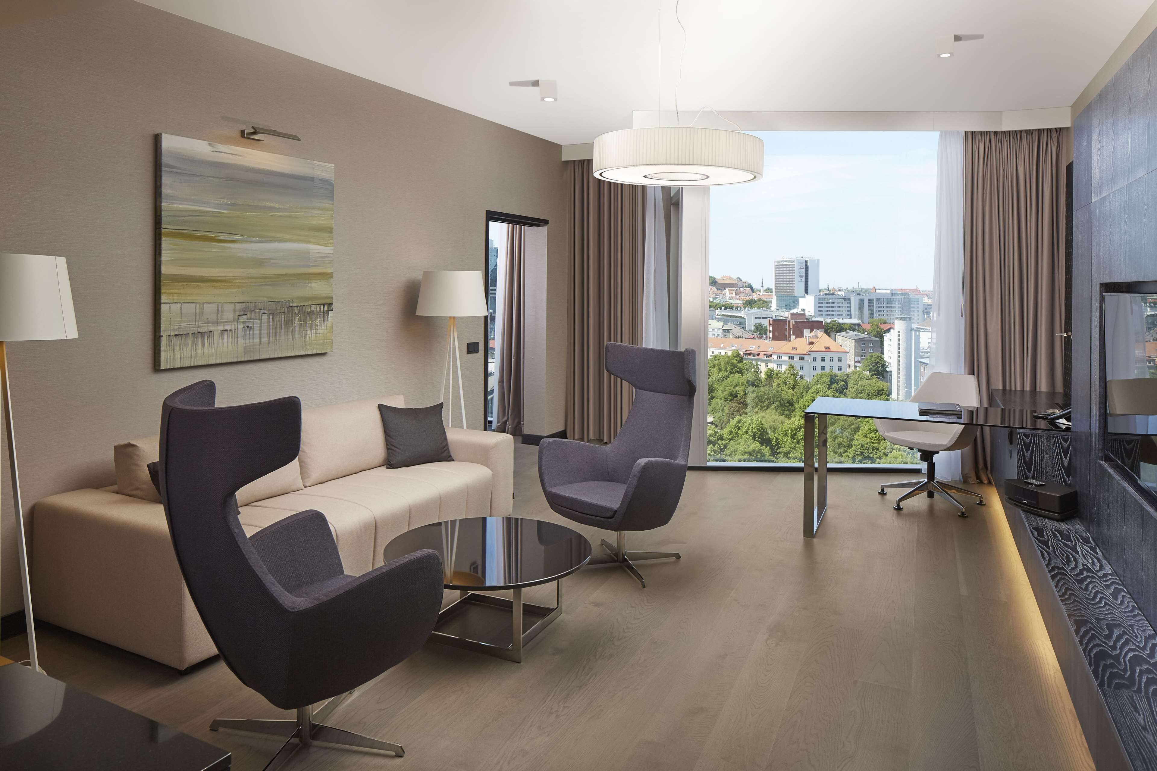 A modern hotel room with a sofa, two chairs, a small table, and a city view through large windows.