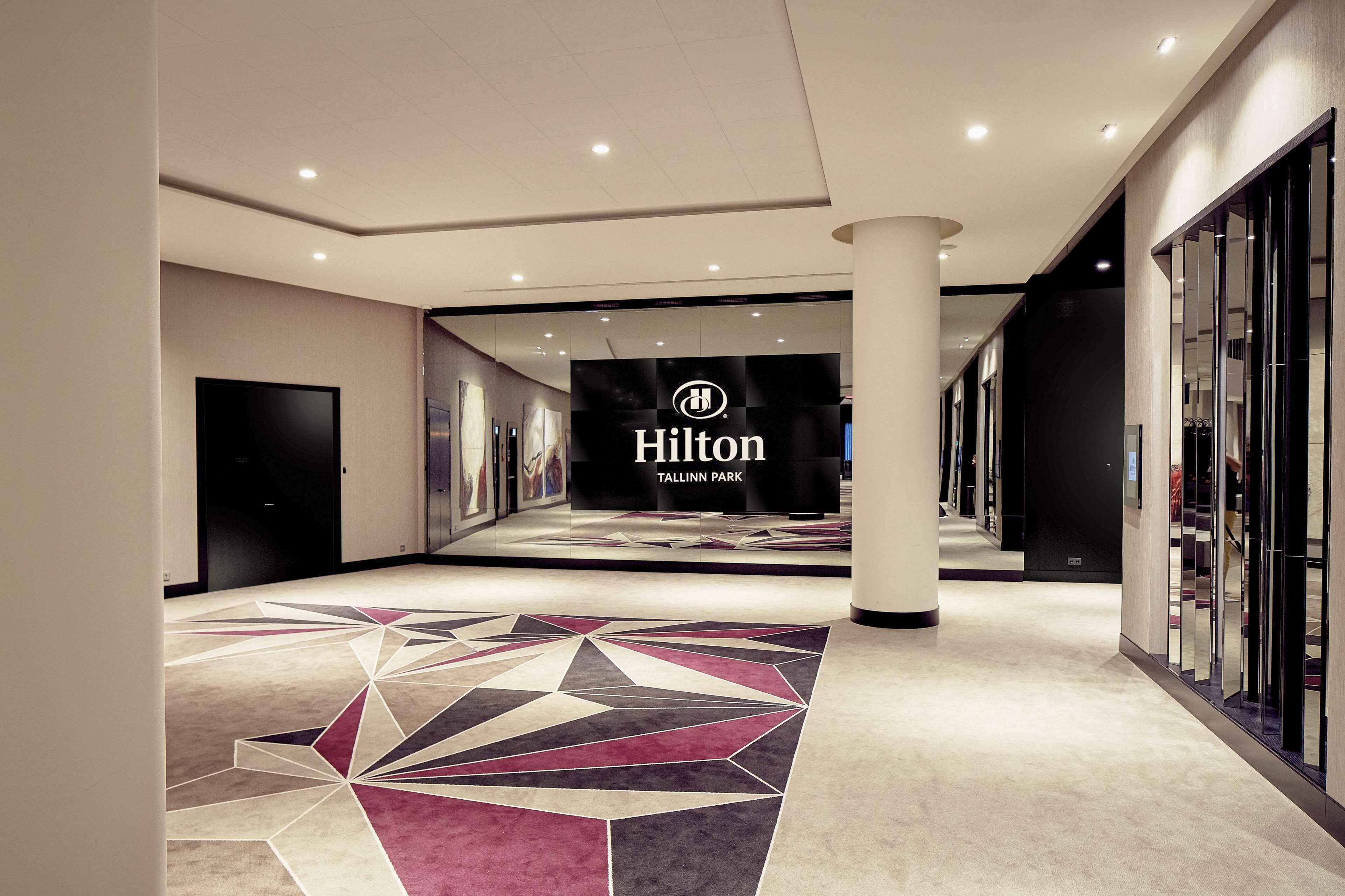 A modern hotel lobby with a geometric carpet design, a prominent Hilton sign, and a spacious corridor with glass doors.