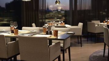 Uma sala de jantar moderna com mesas postas para o jantar, oferecendo vista da cidade por grandes janelas.