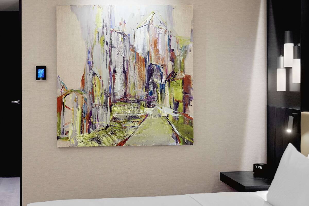 Una camera d'albergo con un grande quadro astratto appeso al muro, un comodino con una sveglia e un letto con lenzuola bianche.