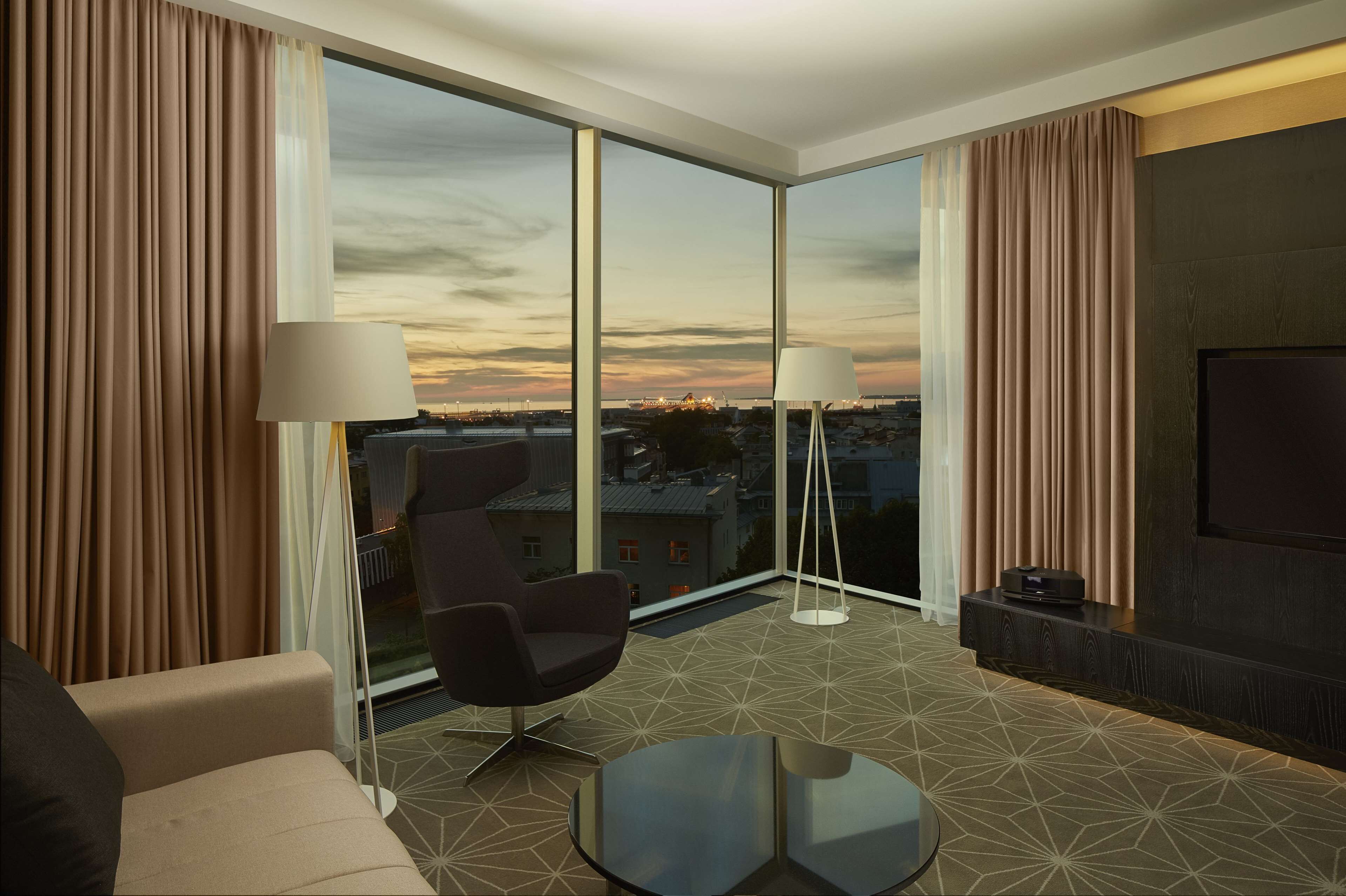 Una habitación de hotel moderna con un ventanal grande, un sofá, una silla, una mesita de centro y un televisor de pantalla plana.
