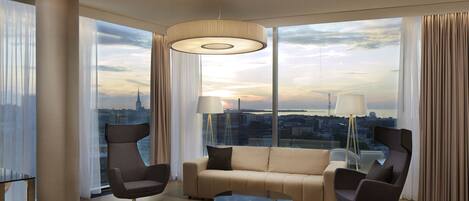 Habitación de hotel moderna con un ventanal grande que ofrece vistas a la ciudad, un sofá, dos sillones y una mesa.