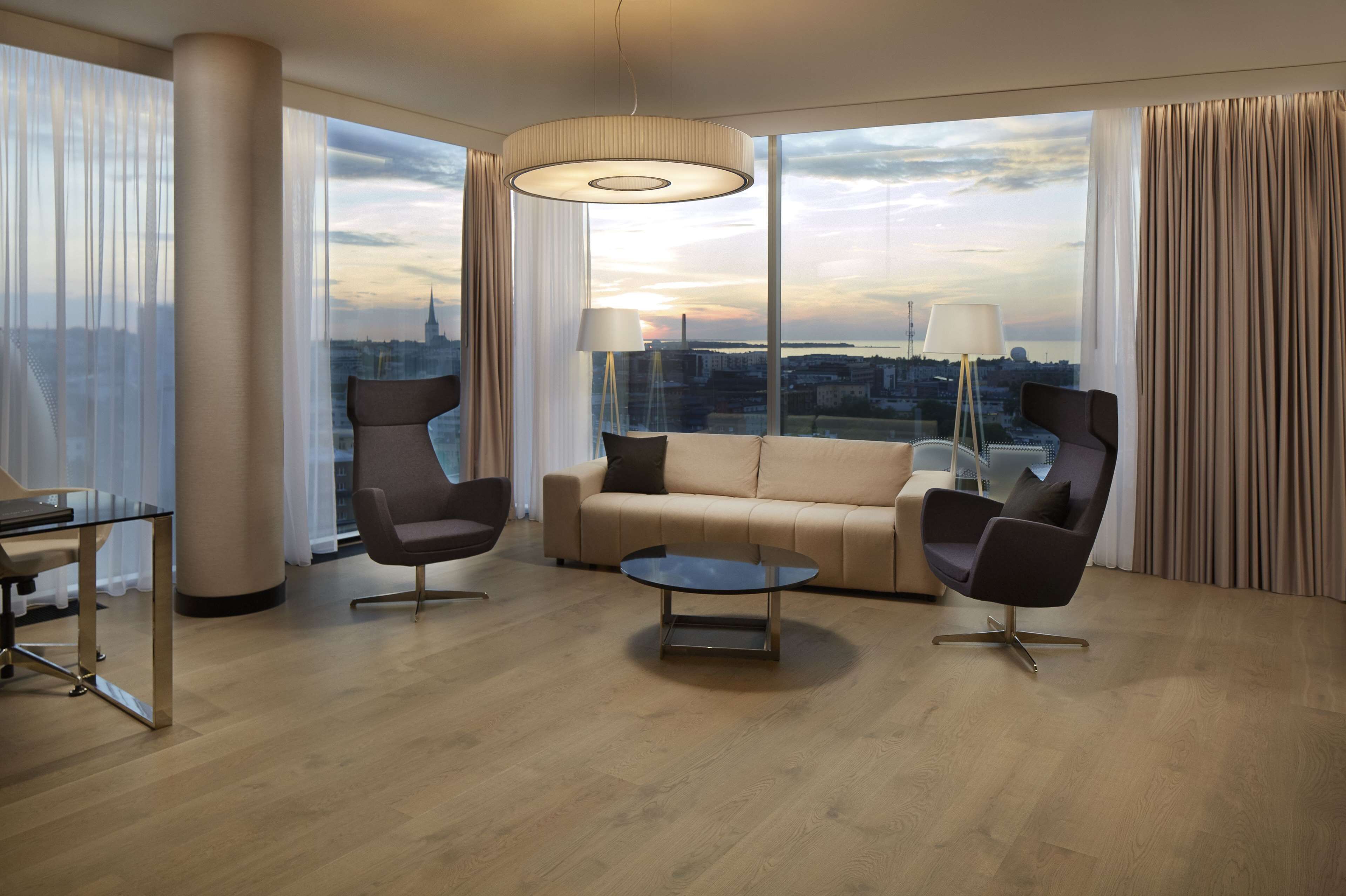 A modern hotel room with a large window offering a city view, a sofa, two armchairs, a coffee table, and a desk with a chair.