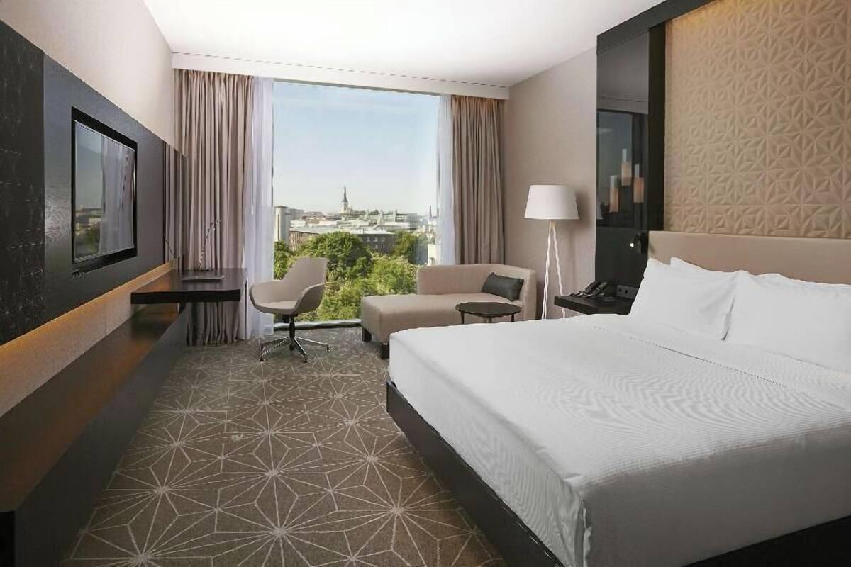 Una camera d'albergo con un letto, una scrivania, una sedia, una lampada e un'ampia finestra vista città.