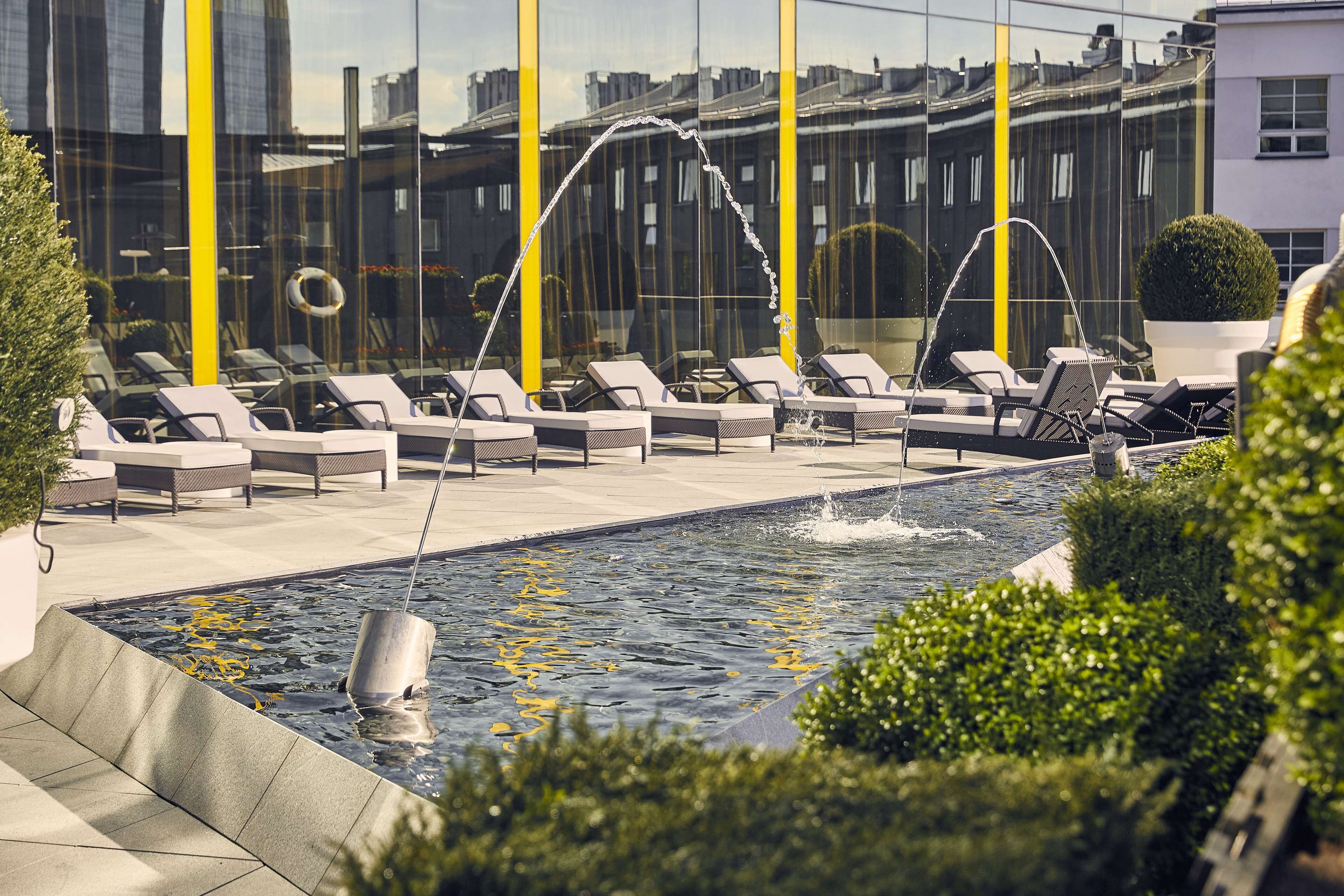 A rooftop pool with water jets, lounge chairs, and a glass barrier reflecting the cityscape.