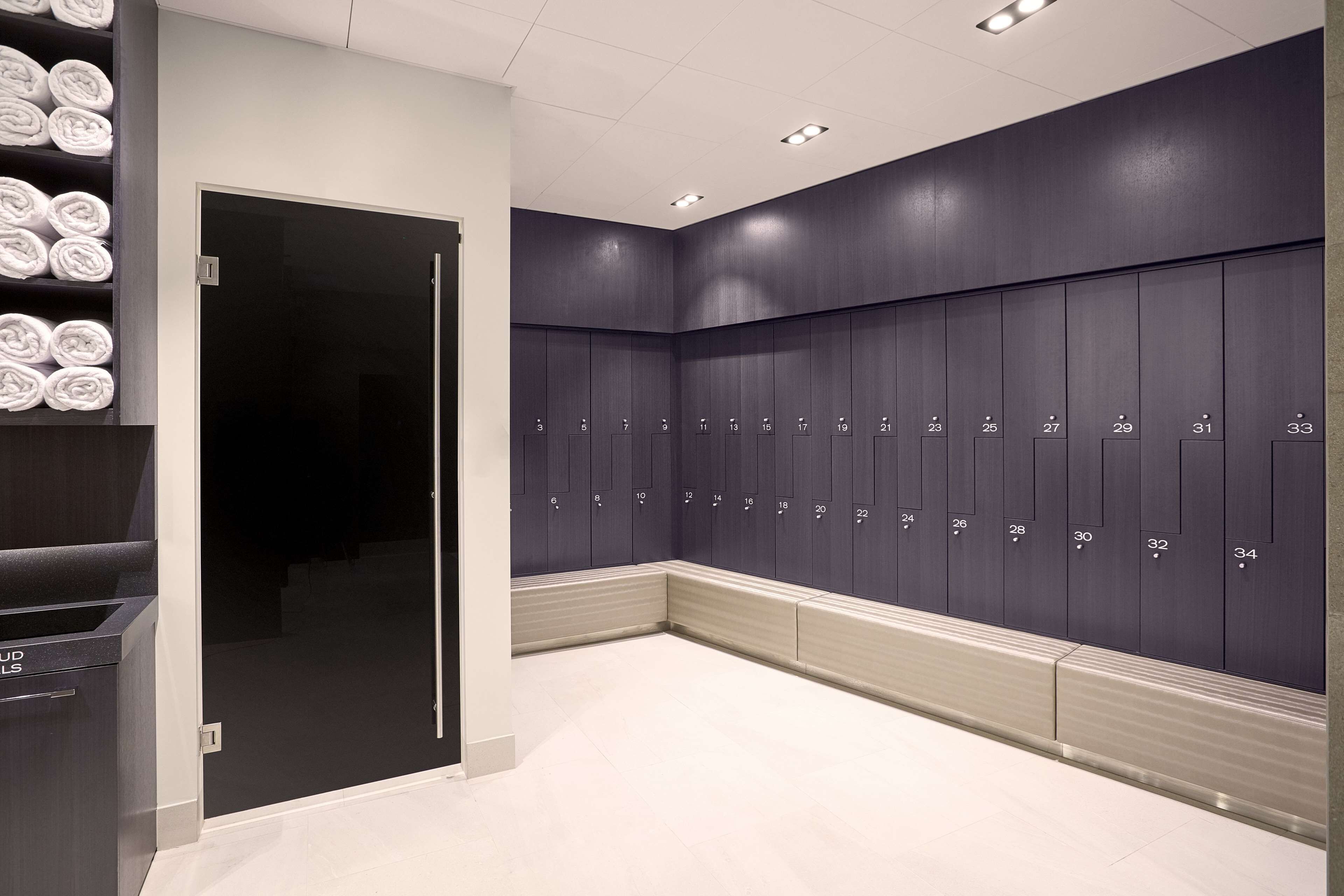 Een moderne kleedkamer met netjes opgeruimde, genummerde kluisjes van 1 tot 34, een glazen deur en een handdoekenrek met gevouwen handdoeken.