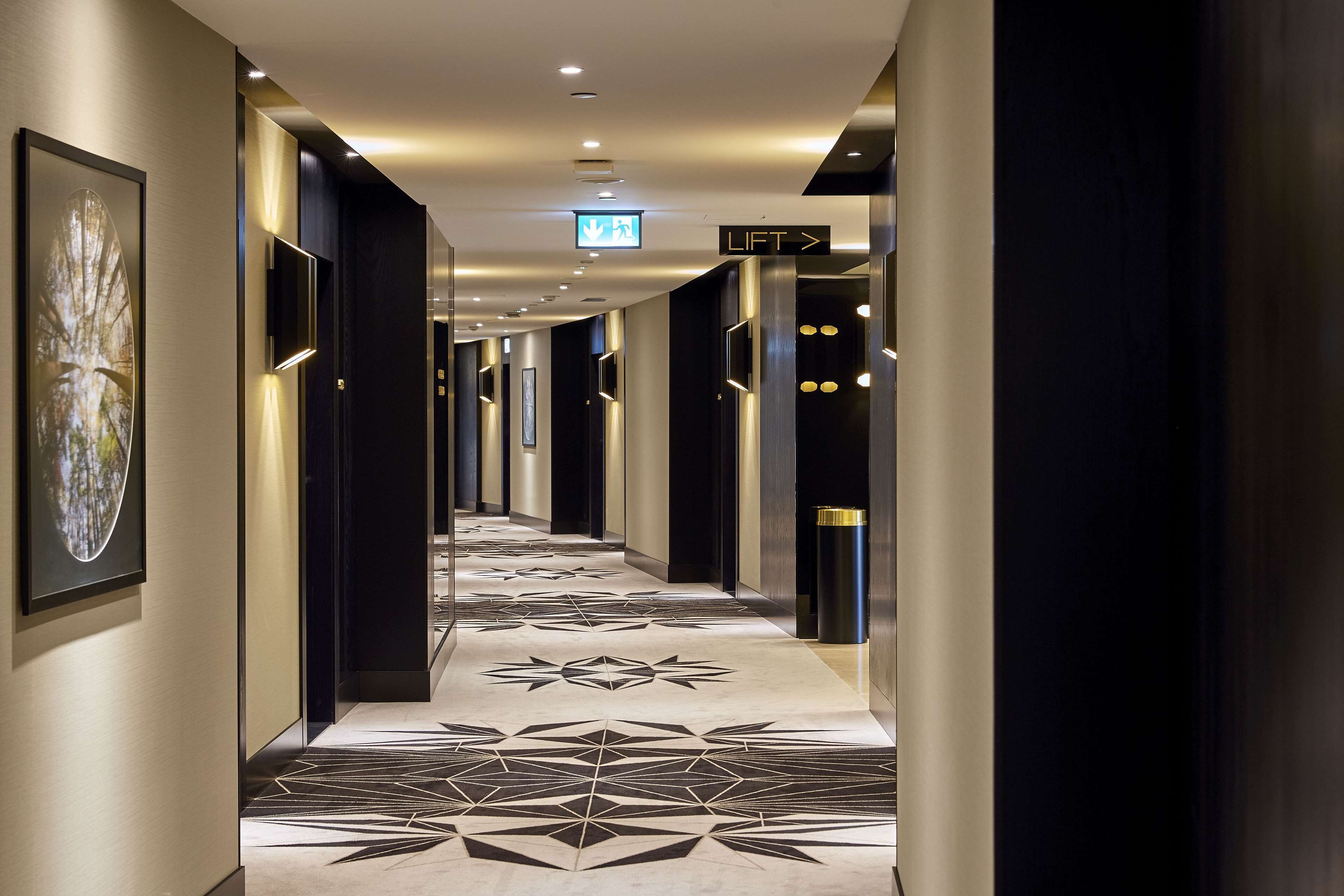 En modern hotellkorridor med ett golv som har ett geometriskt mönster, vägvisningsskyltar och moderna väggmonterade lampor.