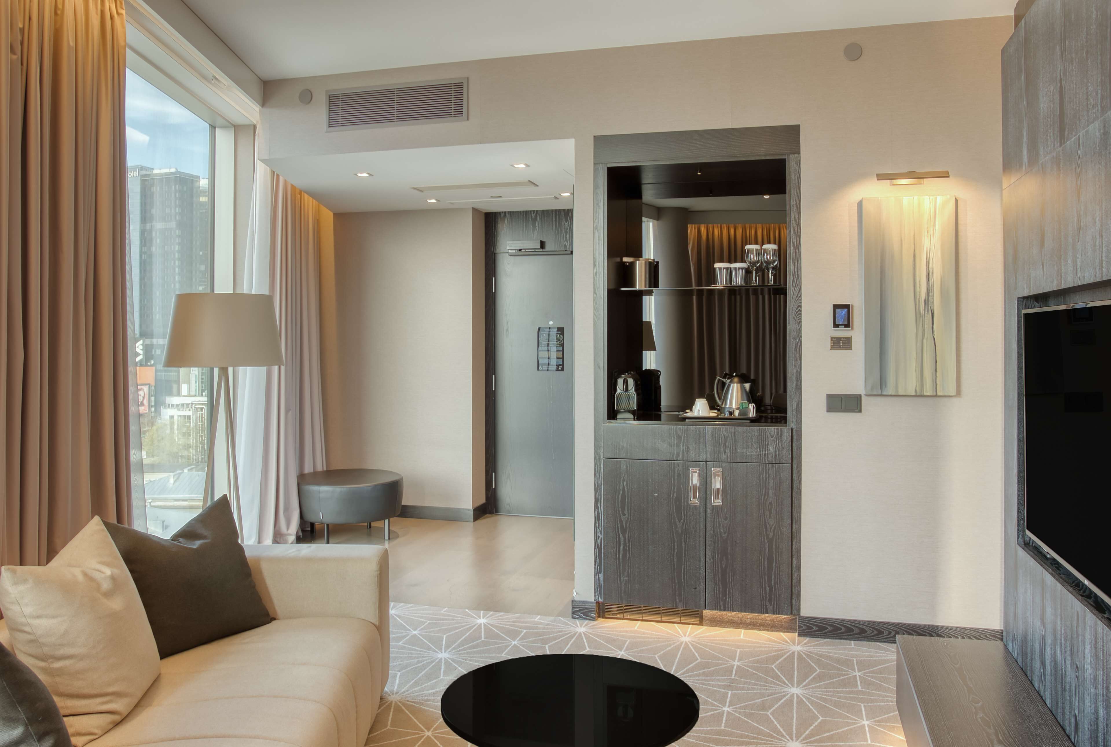 Ein modernes Wohnzimmer mit Sofa, Couchtisch und Fernseher. Es gibt eine Küchenzeile mit Schränken und einer Mikrowelle. Das Zimmer verfügt über große Fenster mit Vorhängen.