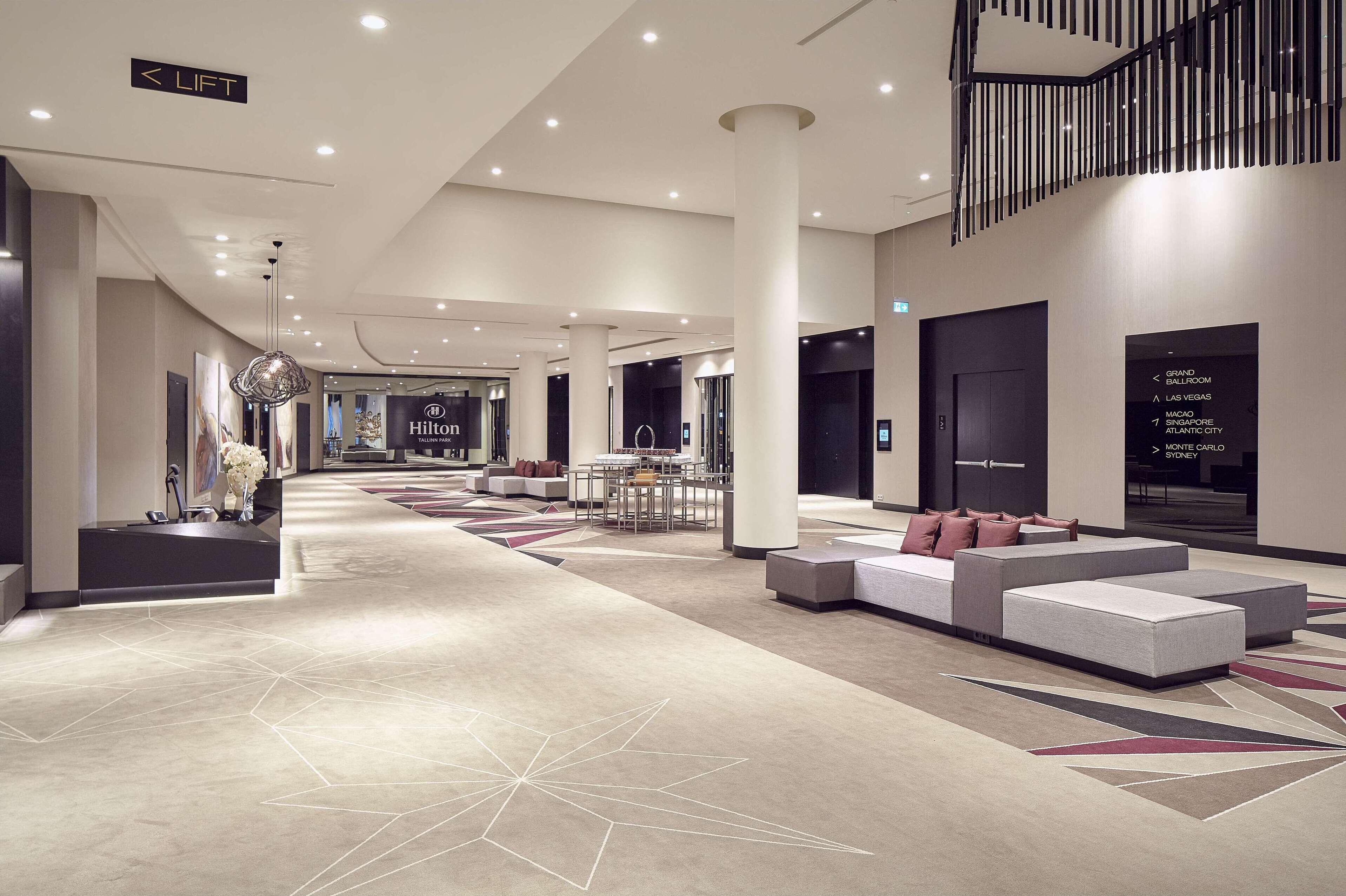 En rummelig hotellobby med et moderne design, med et centralt siddeområde, en receptionsdisk og skilte der viser vej til forskellige områder af hotellet.