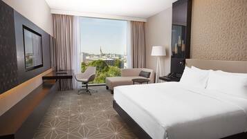A hotel room with a large bed, a desk, a chair, a lamp, and a view of the city.