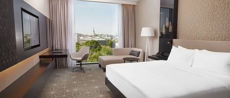 A hotel room with a large bed, a desk, a chair, a lamp, and a view of the city.